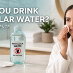 A professional, high-quality blog featured image (1200x628) showing a woman in a white bathrobe gently applying micellar water to her face with a cotton pad in a bright bathroom. In the foreground, a clear bottle of micellar water sits on a marble counter with a prominent "Do Not Drink" warning icon on the label. The text overlay reads: "CAN YOU DRINK MICELLAR WATER? SAFETY & SCIENCE."